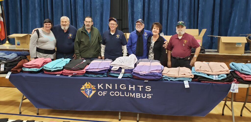 Officers of the Knights of Columbus Council 3660, and two ladies standing in front of a table of coats during the 2023 Coats for Kids event at Central Catholic School.